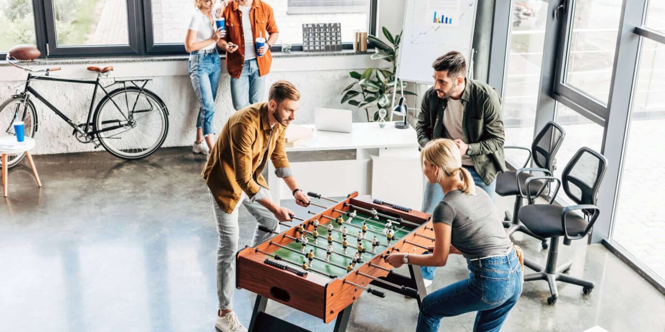 Mitarbeiter spielen Tischfußball im Büro, während Kollegen im Hintergrund entspannt zusammensitzen – ein Beispiel für attraktive Zusatzleistungen im Büro.