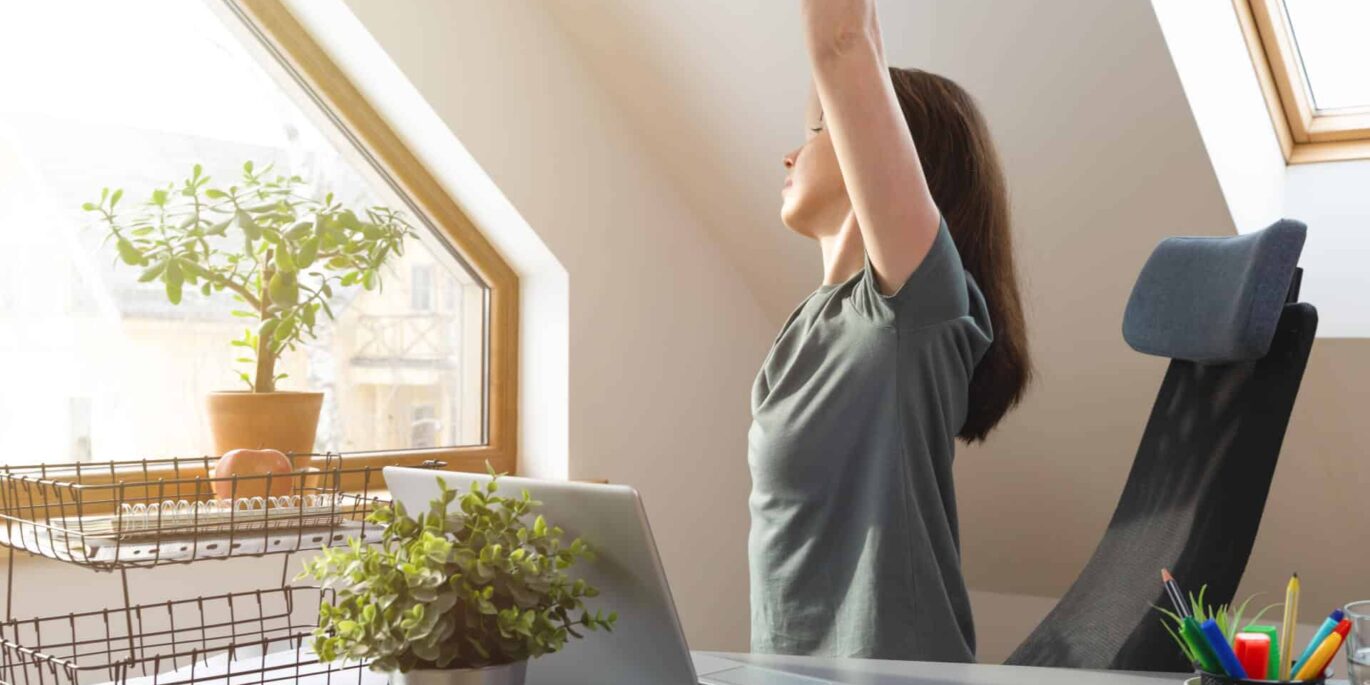 Frau streckt sich entspannt am Arbeitsplatz im Dachgeschoss mit Laptop und Pflanzen auf der Fensterbank – symbolisches Bild für ergonomisches Arbeiten im Homeoffice.