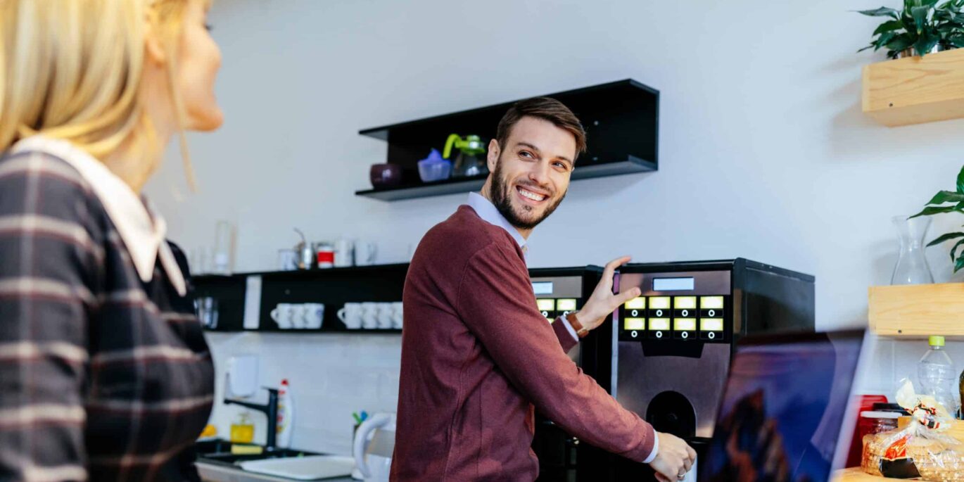Mann lächelt an der Kaffeemaschine und unterhält sich mit Kollegin – Symbolbild für Wohlfühlen im Büro.