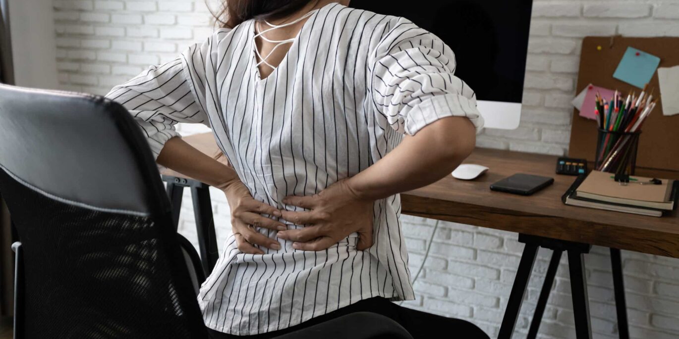 Frau sitzt vor dem PC und fässt sich an den schmerzenden Rücken – Symbolbild für die gesundheitlichen Herausforderungen im Homeoffice.