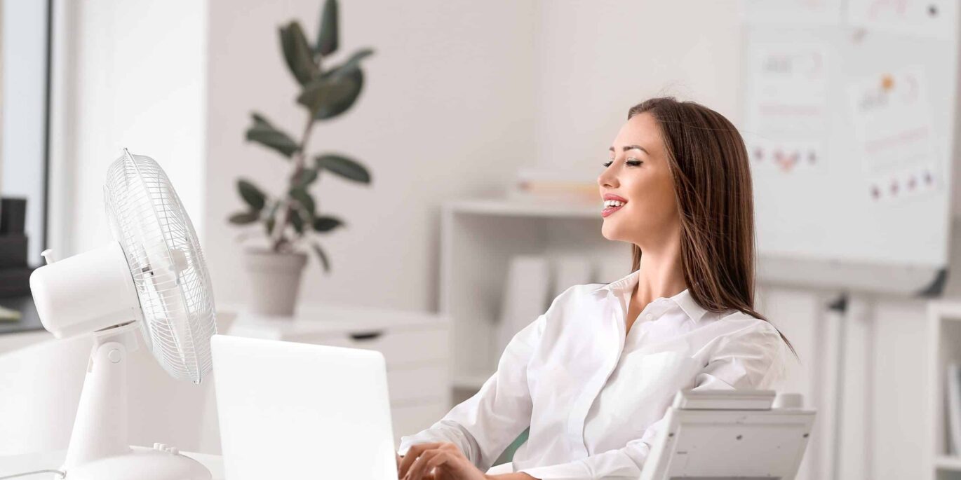 Junge Frau sitzt im Büro vor dem Laptop und genießt die Abkühlung durch einen Ventilator – Sommer im Office.