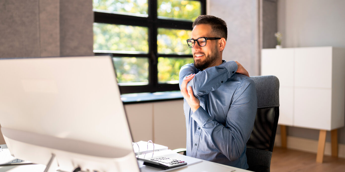 Mann macht Dehnübungen am Arbeitsplatz, um Verspannungen vorzubeugen und schmerzfrei im Büro zu arbeiten.