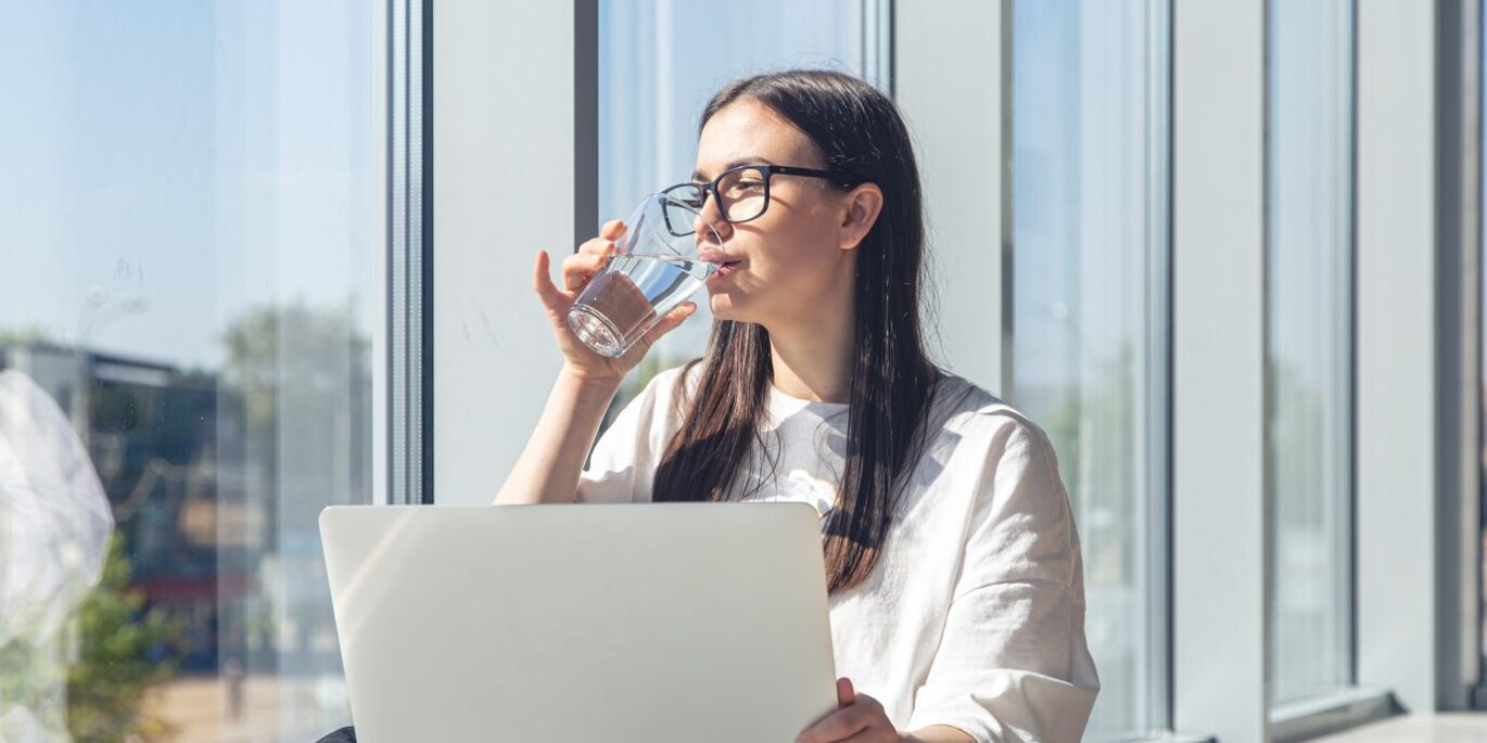 Frau mit Brille trinkt Wasser am Arbeitsplatz vor einem Laptop – Beispiel für gesunde Gewohnheiten im Büro.