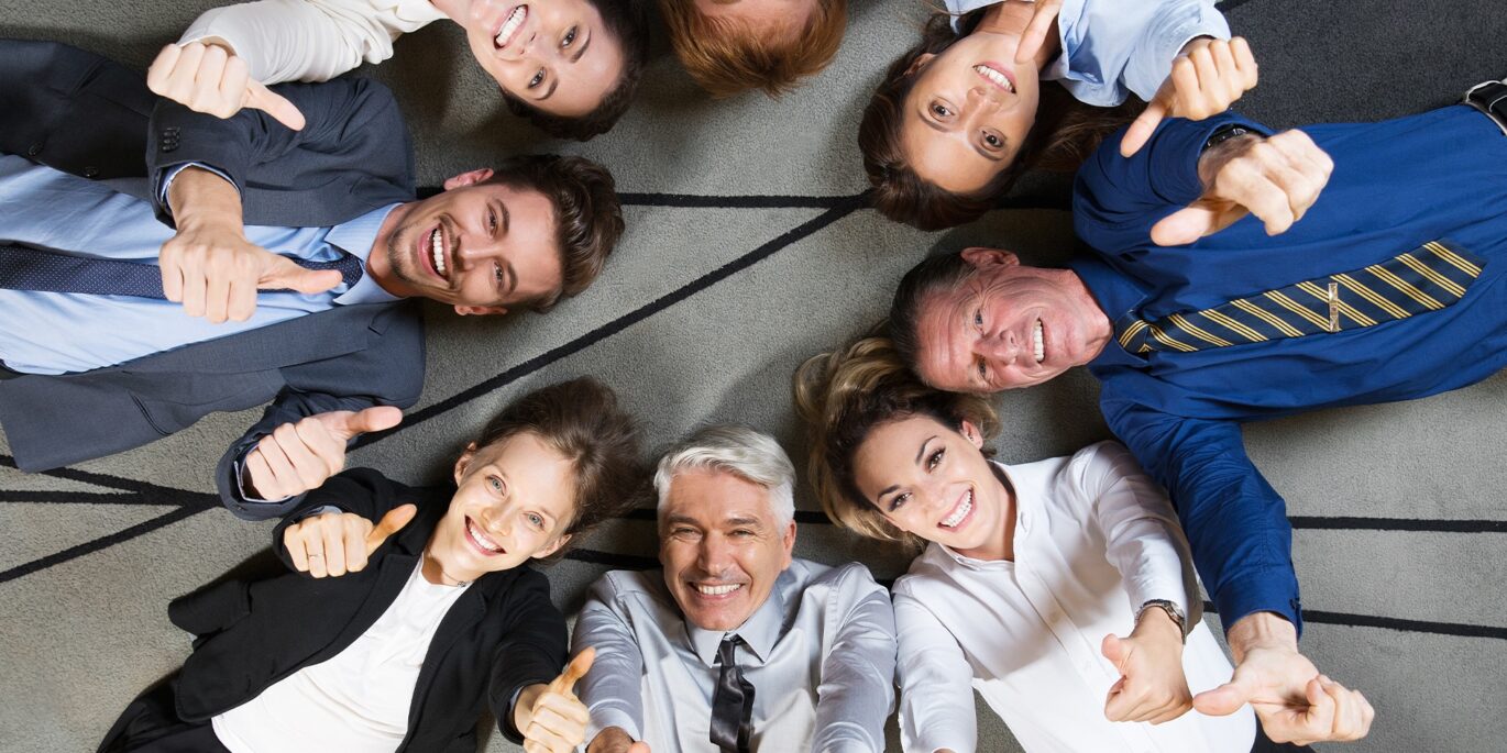 Gruppe von Mitarbeitenden liegt lachend im Kreis auf dem Boden und zeigt Daumen hoch – Symbolbild für verschiedene Mitarbeitertypen.