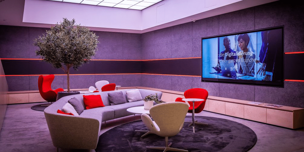 Moderner Pausenraum mit großer Sofaecke, farbigen Akzenten, Lounge-Sesseln und Bildschirm – Gestaltung von Pausenräumen für entspannte Arbeitsatmosphäre.