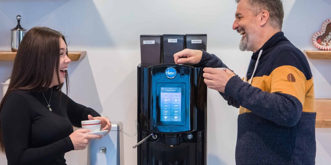 Zwei lachende Personen an einer modernen Kaffeemaschine verdeutlichen, wie Büroeinrichtung und Mitarbeiterzufriedenheit durch gemeinschaftliche Pausen gefördert werden.