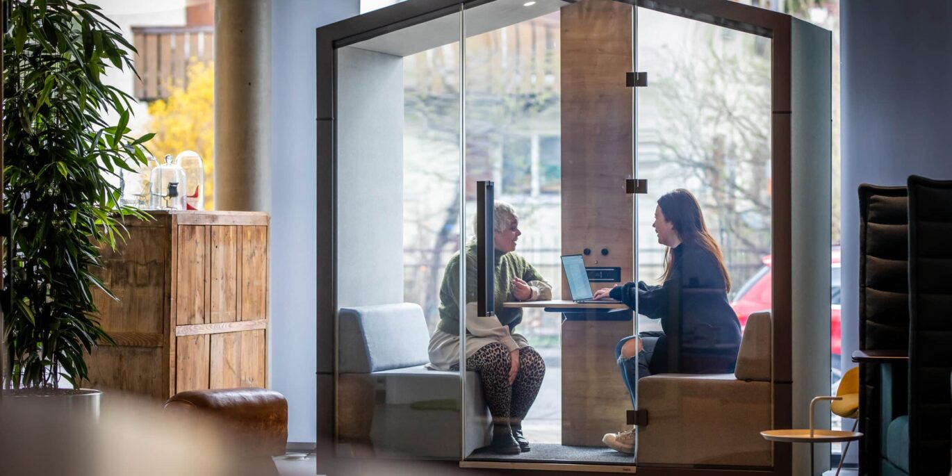 Zwei Frauen arbeiten in einer akustisch abgeschirmten Meetingbox mit Laptop in einem modernen Büro – typische Szene im Kontext von New Work und flexibler Bürogestaltung.