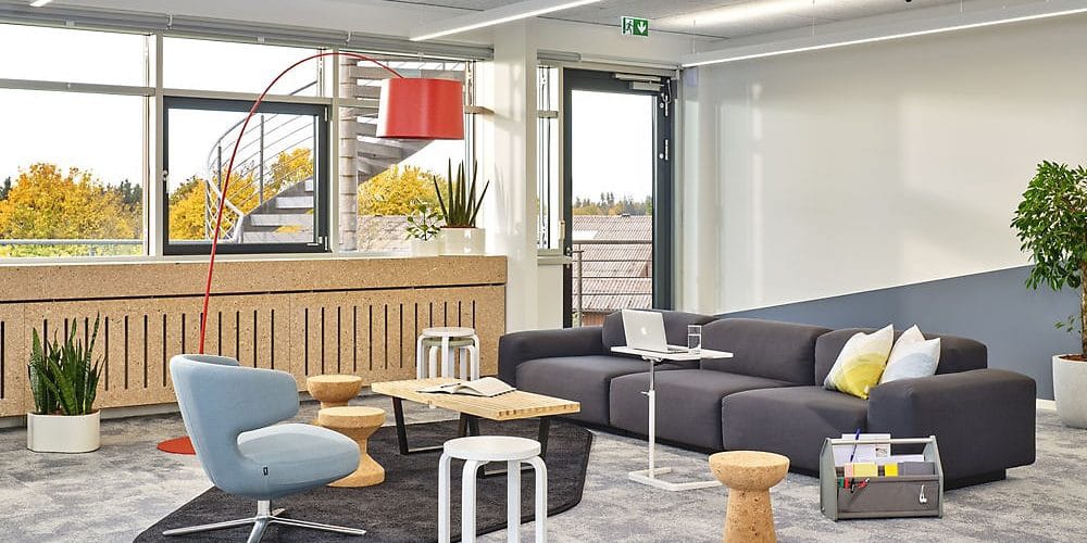 Modern gestaltete Lounge mit grauem Sofa, hellblauem Sessel und roten Akzenten in einer hellen Büroatmosphäre.