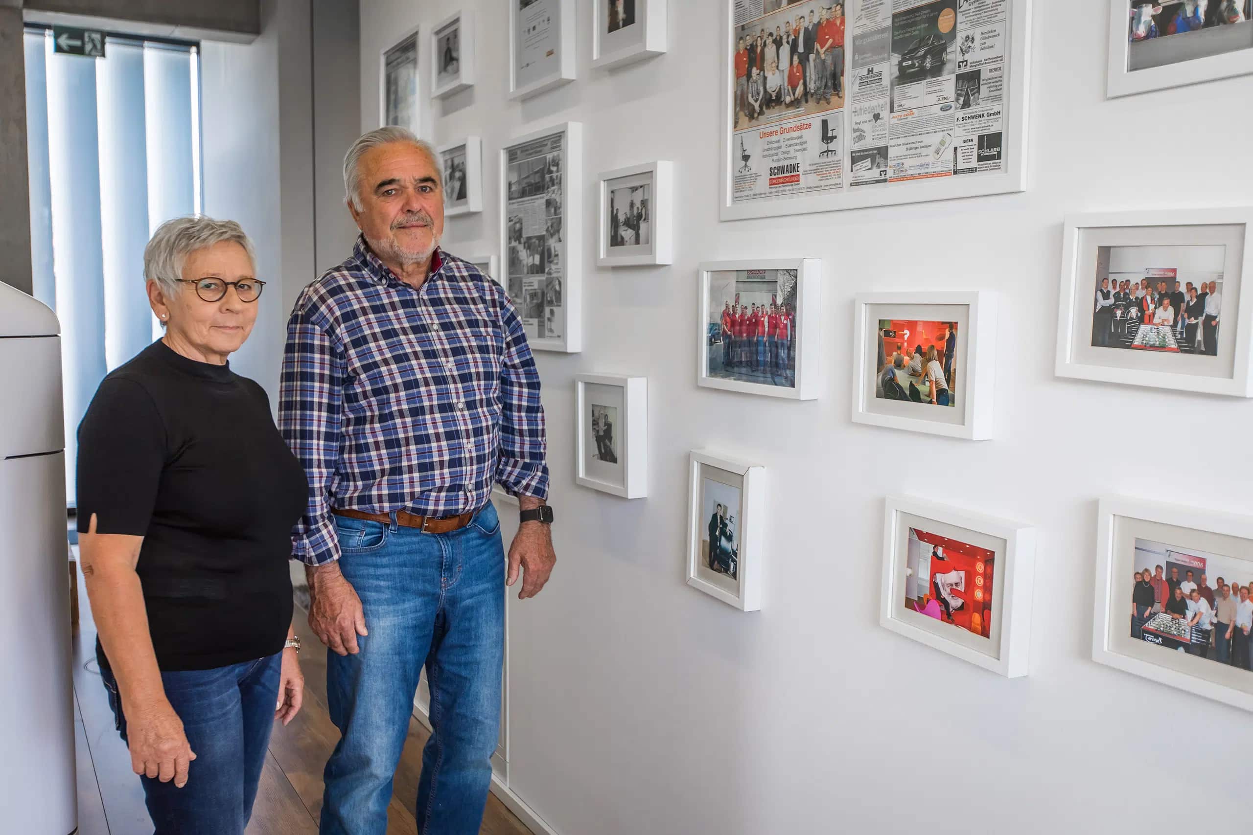 Montageservice-Schwadke-Bueroeinrichtung-Rosenheim Frau und Herr Schwadke stehen vor einer Wand mit gerahmten Fotos und Zeitungsartikeln – Symbolbild für den Montageservice