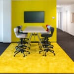 Moderner Meetingbereich im Büro mit hoher Tischlösung, Drehstühlen und gelbem Teppich als klar abgegrenzte Kommunikationszone – Beispiel für Flächenzonierung im Open Space.