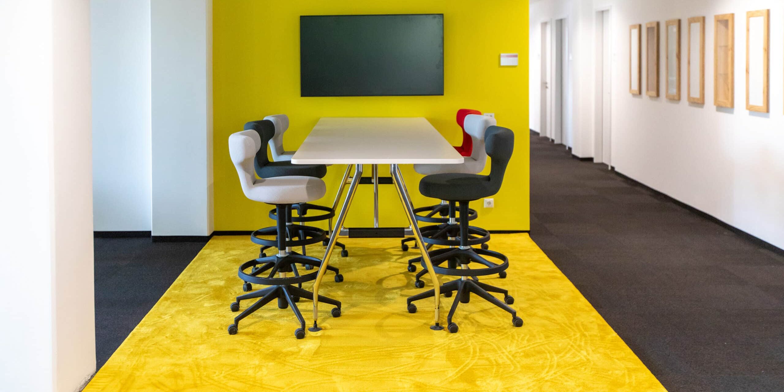 Flächenzonierung Schwadke Büroeinrichtungen Moderner Meetingbereich im Büro mit hoher Tischlösung, Drehstühlen und gelbem Teppich als klar abgegrenzte Kommunikationszone – Beispiel für Flächenzonierung im Open Space.