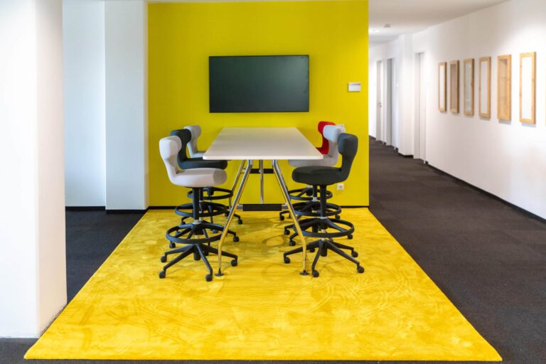 Flächenzonierung Schwadke Büroeinrichtungen Moderner Meetingbereich im Büro mit hoher Tischlösung, Drehstühlen und gelbem Teppich als klar abgegrenzte Kommunikationszone – Beispiel für Flächenzonierung im Open Space.