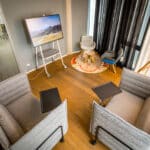 Gemütlicher Besprechungsraum mit zwei gepolsterten Lounge-Sofas mit integrierten Ablageflächen, einem mobilen Bildschirm auf Rollständer und einer kleinen Sitzecke mit Sesseln und Teppich im Hintergrund; warme Holzoptik und wohnliche Gestaltung im Resimercial Design Stil