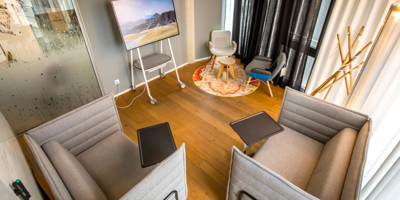 Gemütlicher Besprechungsraum mit zwei gepolsterten Lounge-Sofas mit integrierten Ablageflächen, einem mobilen Bildschirm auf Rollständer und einer kleinen Sitzecke mit Sesseln und Teppich im Hintergrund; warme Holzoptik und wohnliche Gestaltung im Resimercial Design Stil