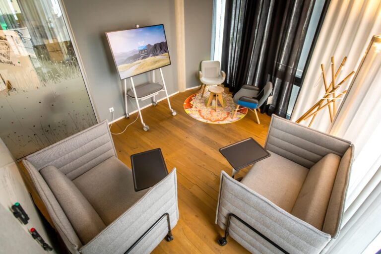 Gemütlicher Besprechungsraum mit zwei gepolsterten Lounge-Sofas mit integrierten Ablageflächen, einem mobilen Bildschirm auf Rollständer und einer kleinen Sitzecke mit Sesseln und Teppich im Hintergrund; warme Holzoptik und wohnliche Gestaltung im Resimercial Design Stil